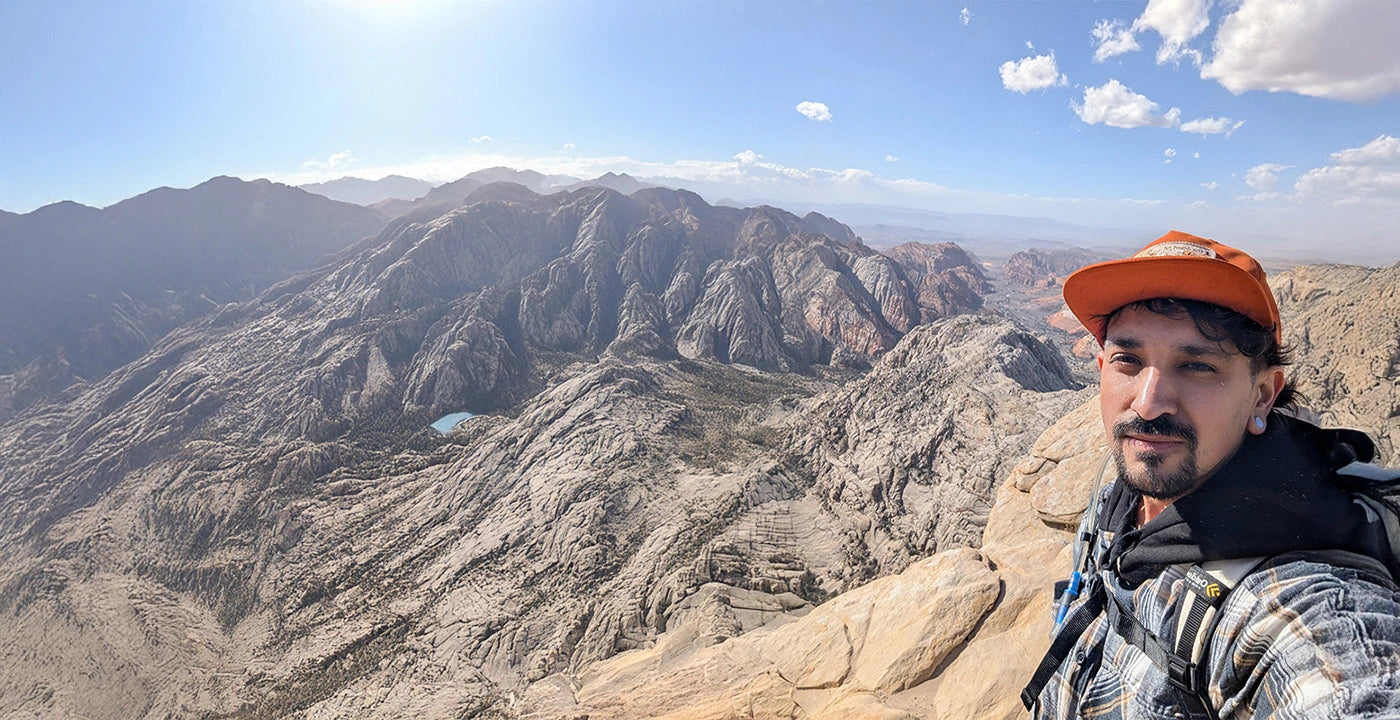 Man taking a selfie on a mountain peak with a scenic view of mountains and sky.